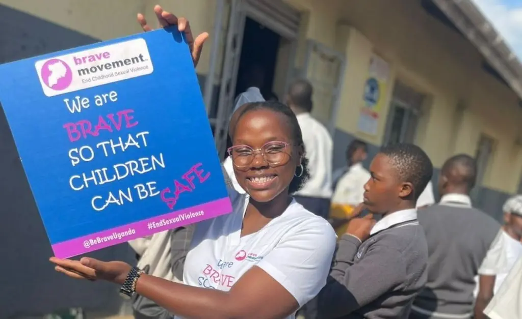 Image shows a smiling woman holding a sign that says "We are brave so that children can be safe" with the Brave Movement logo. Behind her are other people, likely children, also visible in the image.