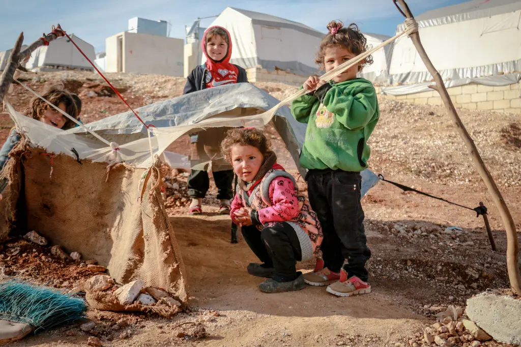Three young children playing in a refugee camp, makeshift tents and shelters in the background, children wearing colorful clothing.