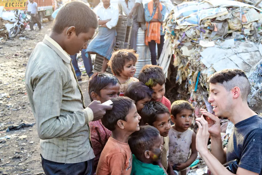 A group of children, some adults, and a man surrounded by debris in what appears to be an urban slum or impoverished area.