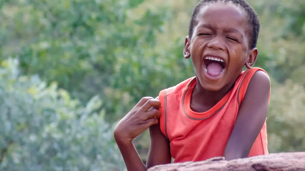 A young person with dark skin wearing a red shirt laughing joyfully in an outdoor setting with greenery in the background.