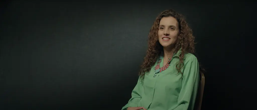 Smiling person with curly brown hair wearing a green shirt on a dark background.