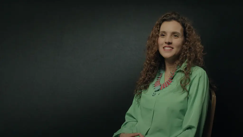 Smiling person with curly brown hair wearing a green shirt on a dark background.