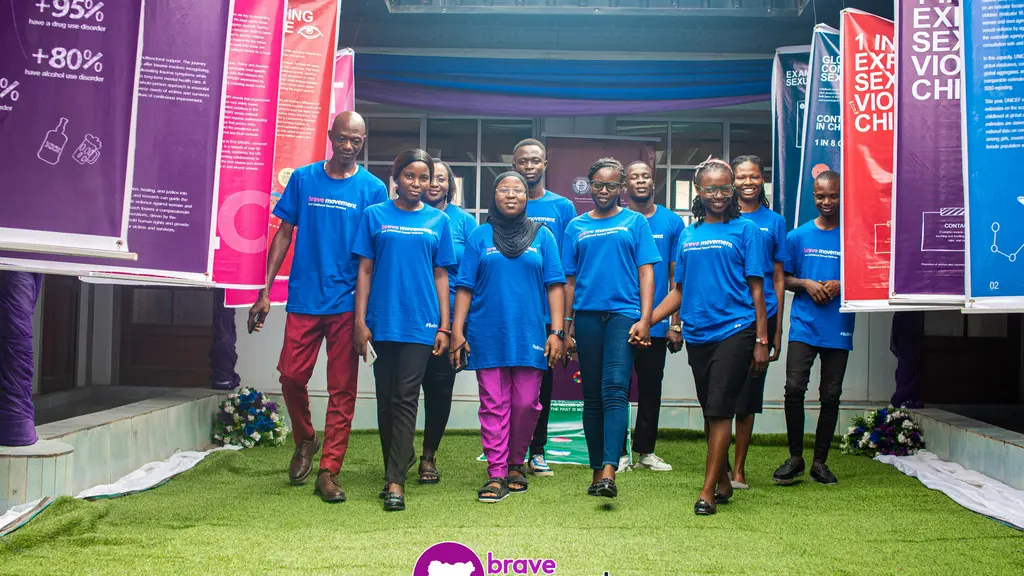 Group of Ugandan children wearing blue tops standing together outdoors, with Brave Movement logo visible.