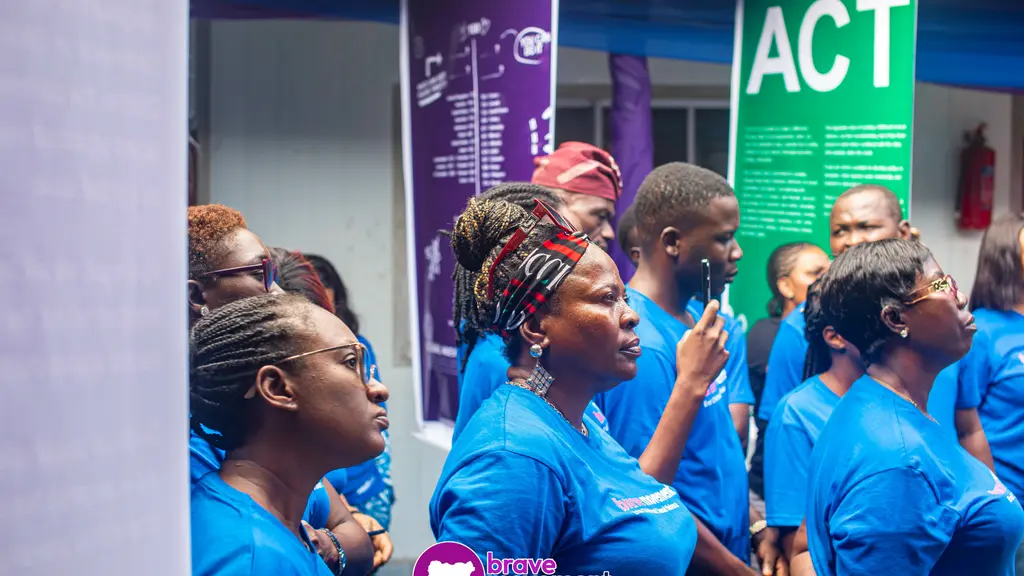 Diverse group of people in blue uniforms, standing and conversing in front of a green 'ACT' sign on a purple wall.