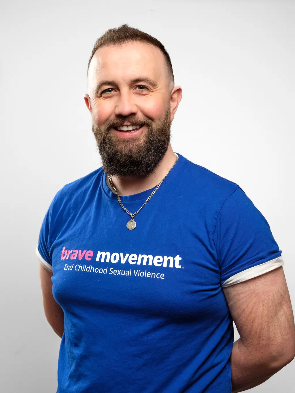 A smiling man with a beard wearing a blue t-shirt with the text "brave movement. End Childhood Sexual Violence".
