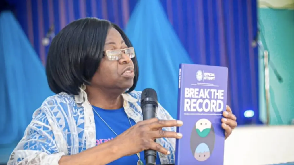 A woman speaking into a microphone while holding a book titled "Break the Record" on a podium against a blue and purple backdrop.