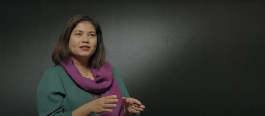 A young woman with long brown hair wearing a green top and purple scarf, standing against a dark background.