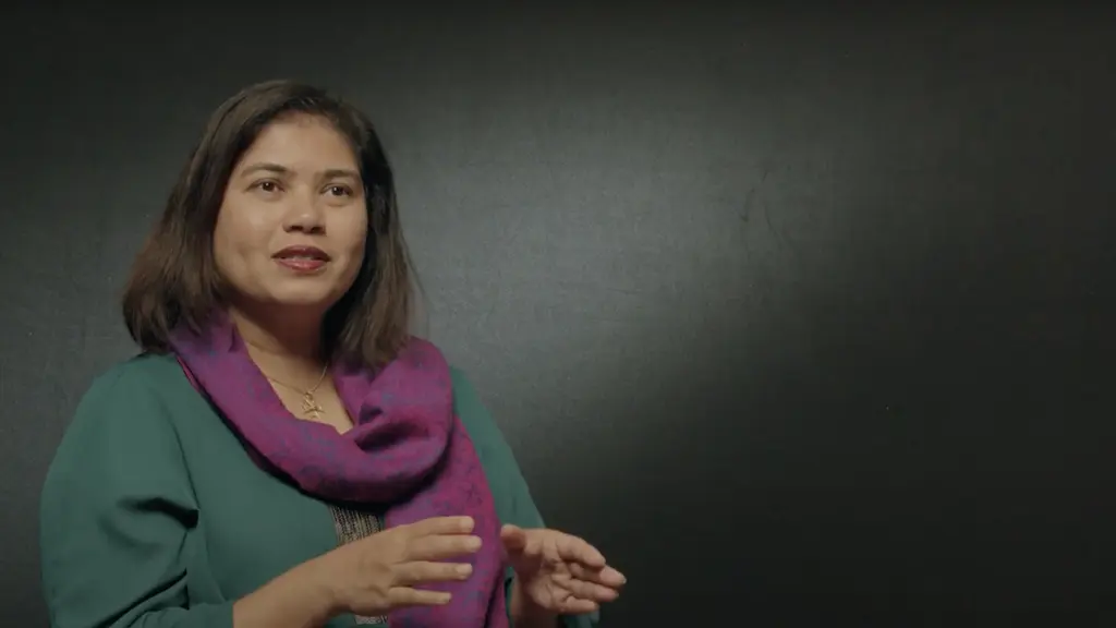 A young woman with long brown hair wearing a green top and purple scarf, standing against a dark background.