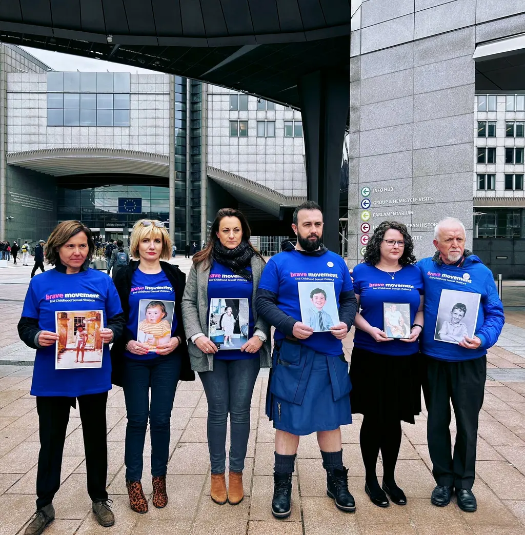 Six individuals holding framed photographs of missing children, standing in front of a modern glass and steel building complex.