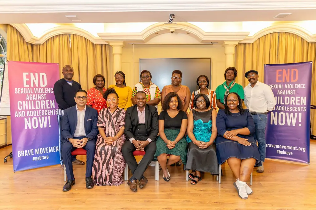 Group of adults, diverse in gender and ethnicity, holding signs that read "End sexual violence against children and adolescents NOW!" and "BRAVE MOVEMENT", seated together in a formal setting with curtains and decor.