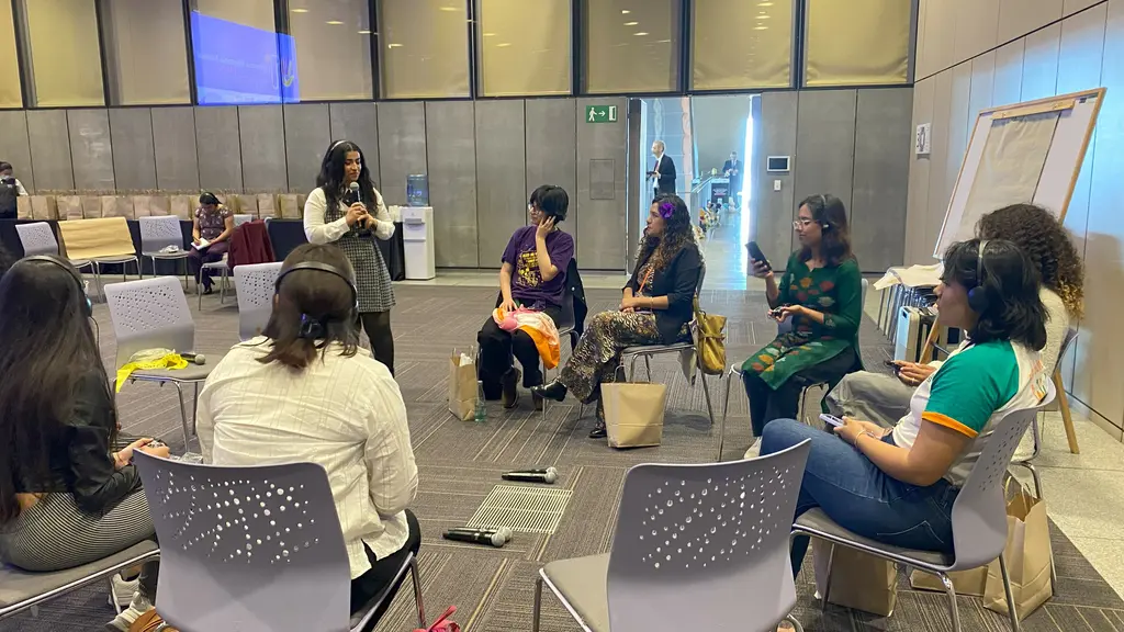 A group of people seated in chairs, having a discussion or meeting in an indoor space. The room has modern furnishings and decor, with a mix of chairs and tables.