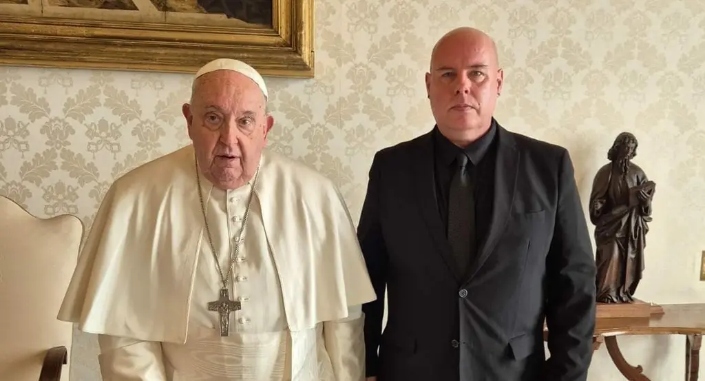 Two elderly men, one in a papal robe and the other in a suit, standing in a room with ornate furnishings and artwork.
