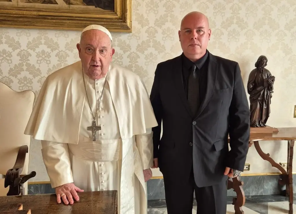 Two elderly men, one in a papal robe and the other in a suit, standing in a room with ornate furnishings and artwork.
