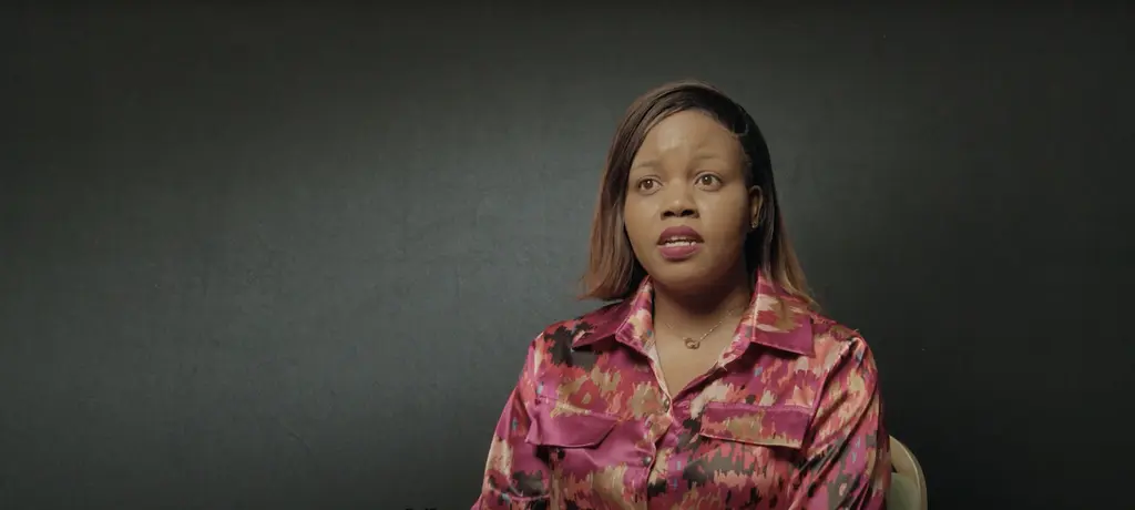 A woman with dark skin and long straight hair, wearing a bright pink patterned shirt, looking directly at the camera.
