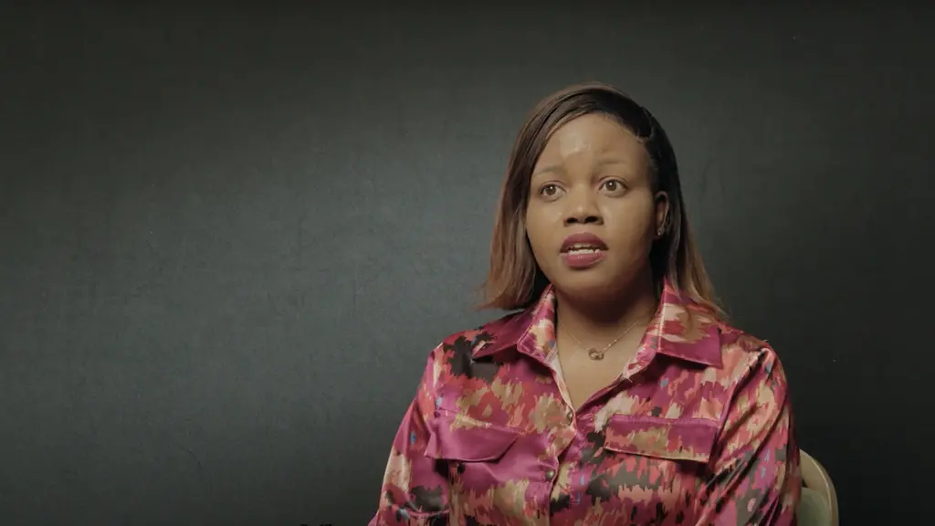 A woman with dark skin and long straight hair, wearing a bright pink patterned shirt, looking directly at the camera.