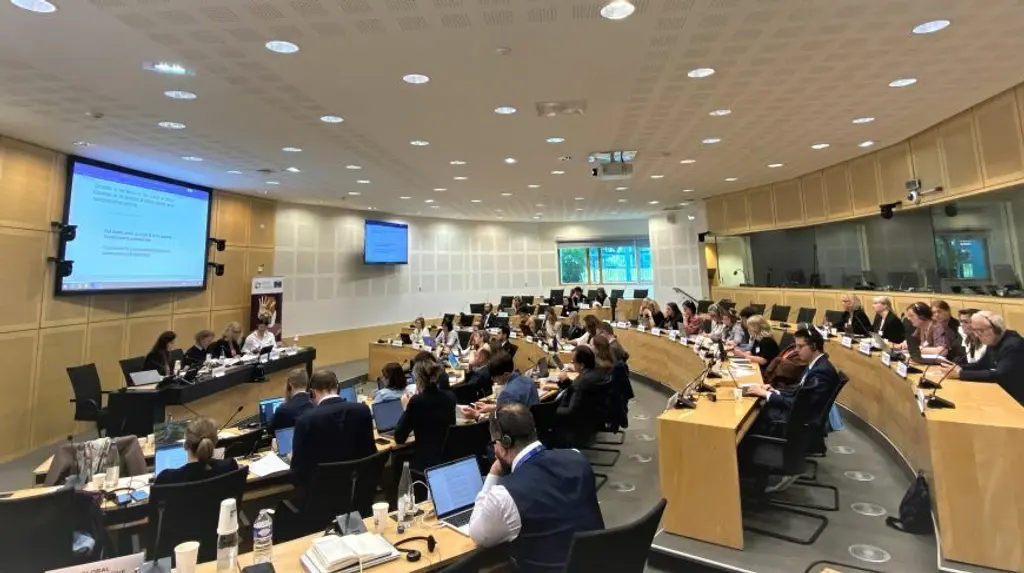 Crowded meeting room with many people seated around a large conference table, with a projection screen at the front.