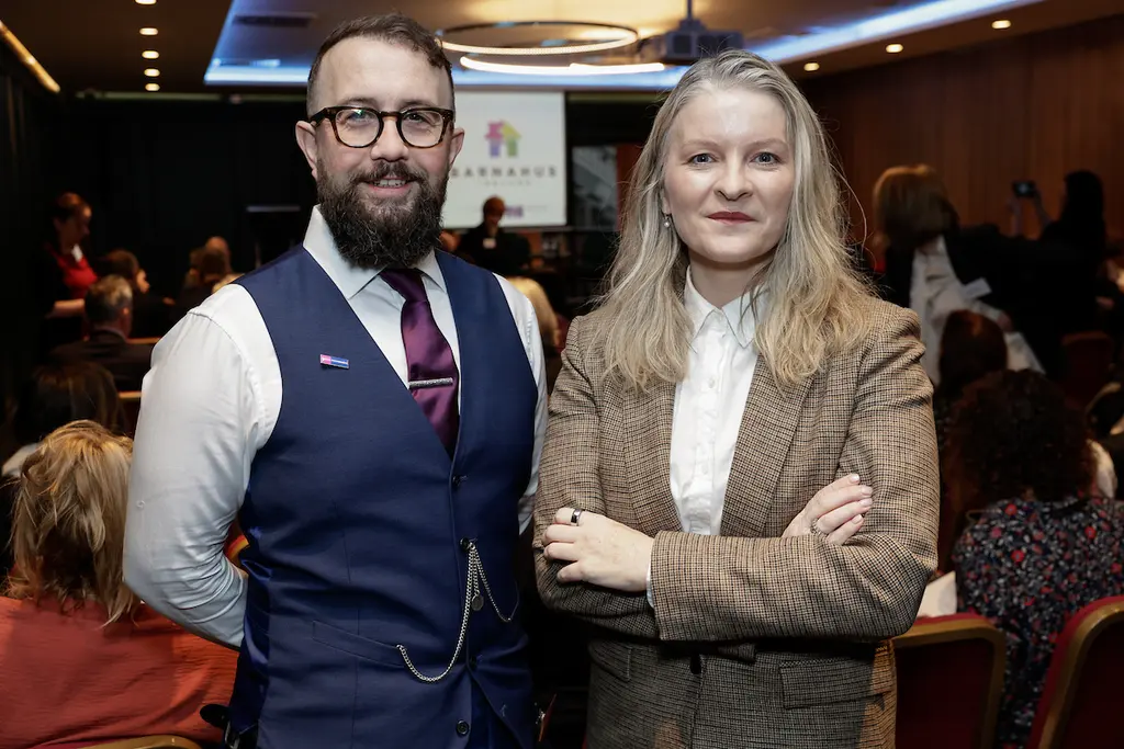 Two people, a man and a woman, standing together in a formal setting. The man is wearing a waistcoat and tie, and the woman is wearing a blazer and blouse.
