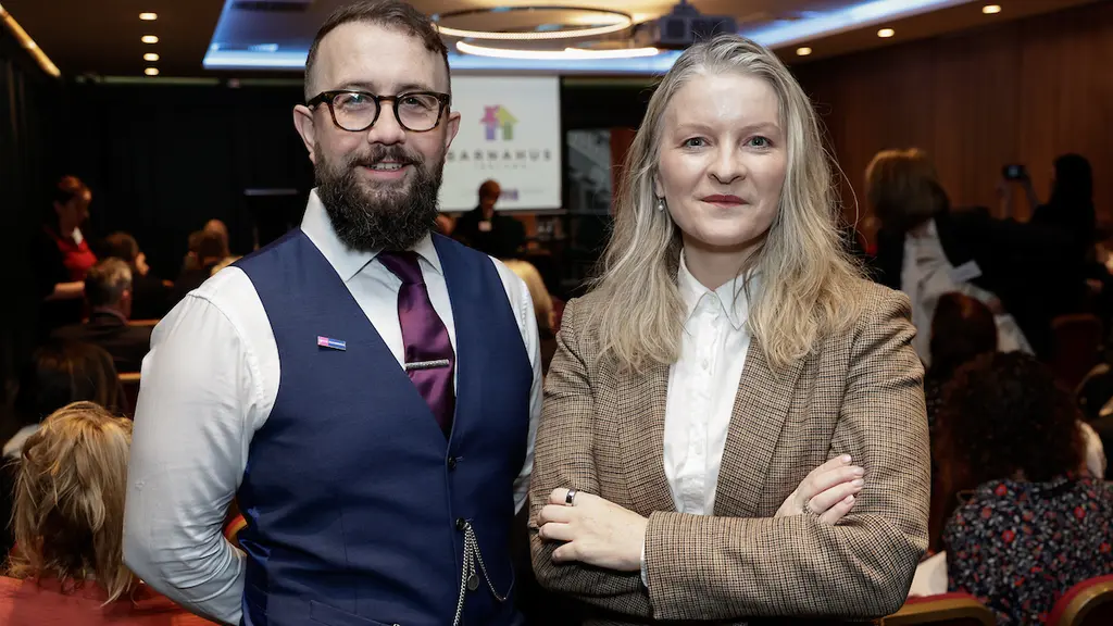 Two people, a man and a woman, standing together in a formal setting. The man is wearing a waistcoat and tie, and the woman is wearing a blazer and blouse.