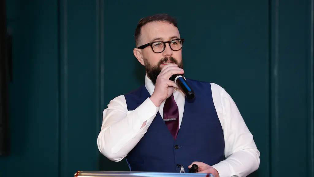 A bearded man in glasses and a suit speaking into a microphone at a podium.