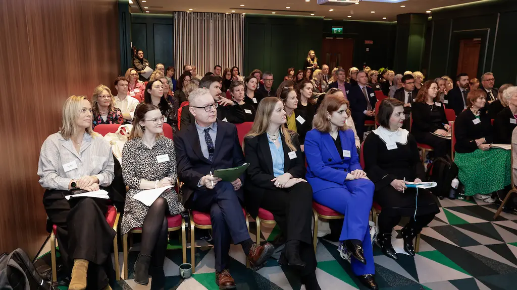 Audience in an auditorium listening to a presentation, seated in rows on chairs with a stage and curtain visible.