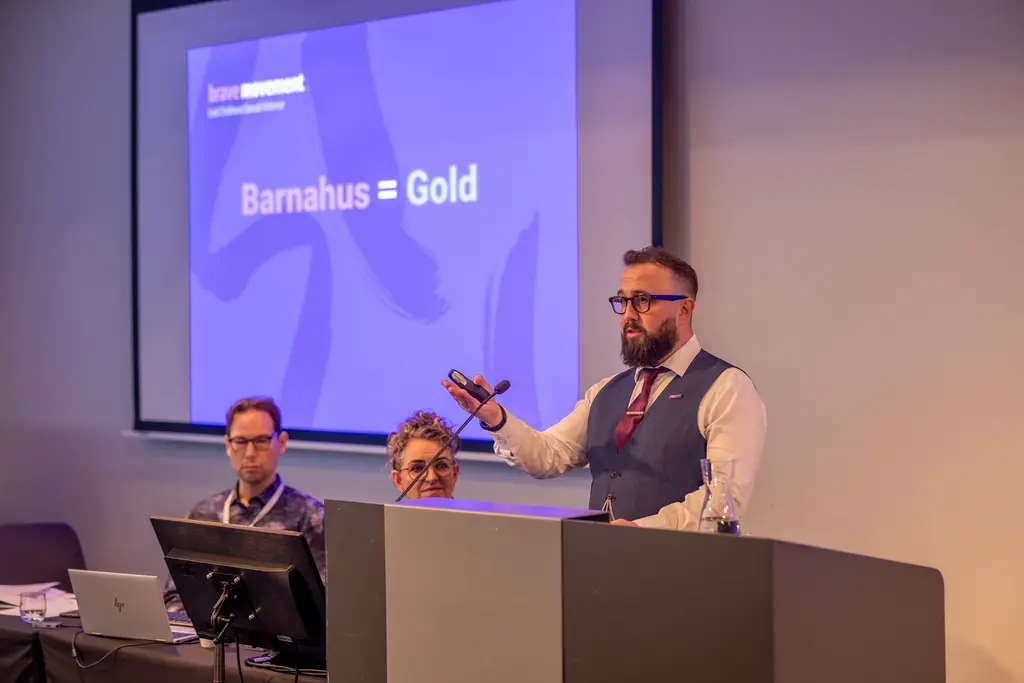 Bearded man speaking at podium, presenting on screen showing "Barnahus = Gold"; two other men seated behind him.