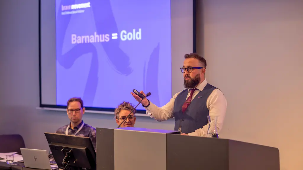 Bearded man speaking at podium, presenting on screen showing "Barnahus = Gold"; two other men seated behind him.