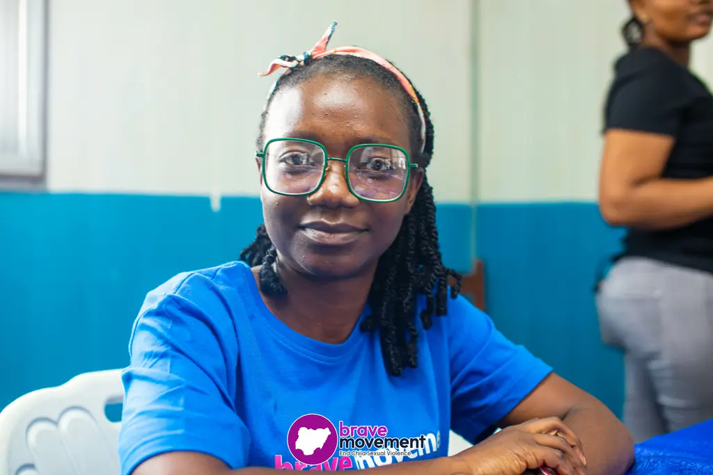 Young woman in blue t-shirt and glasses, with braided hairstyle, seated at a desk.