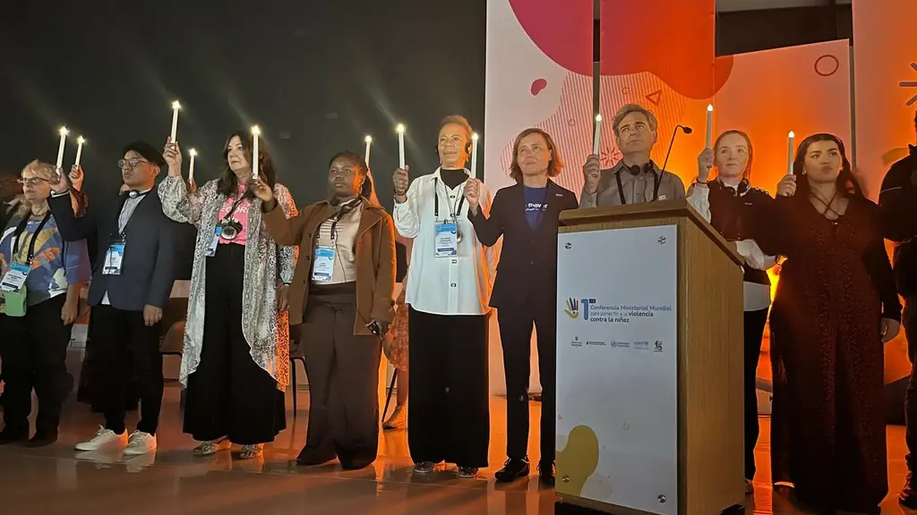People standing on stage in formal and casual attire, with colorful lighting and banners in the background.