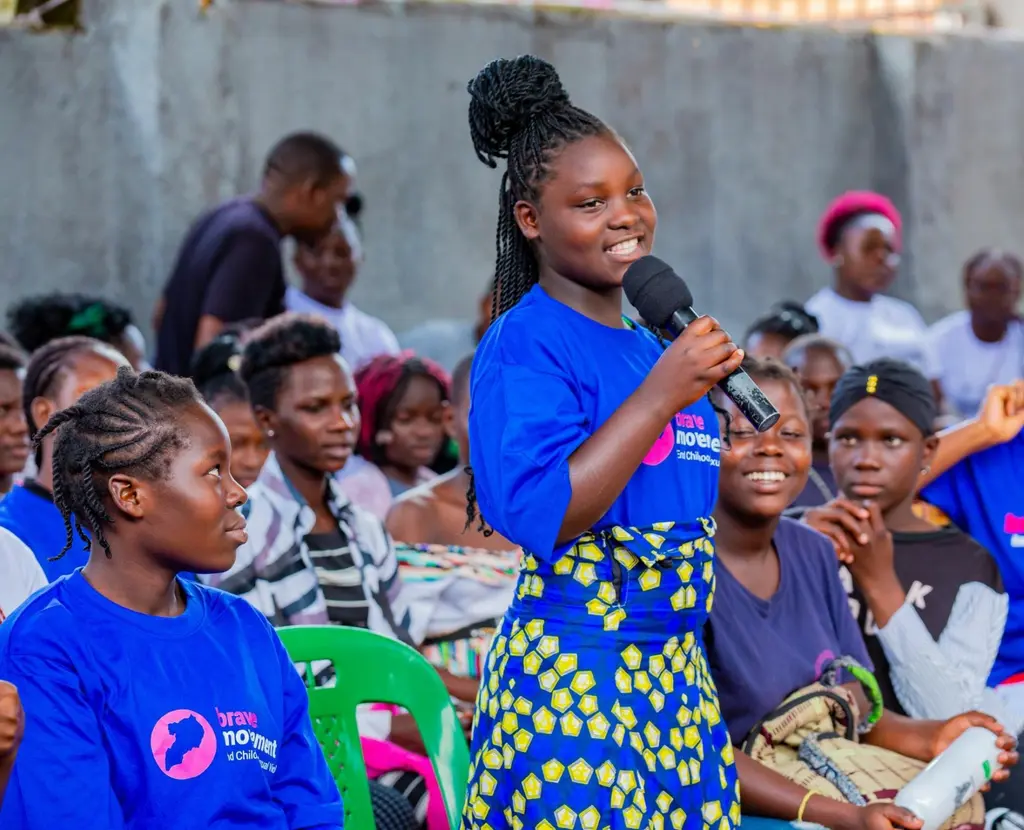 Young girl speaking into microphone surrounded by members of Brave Movement, wearing blue Brave t-shirt.
