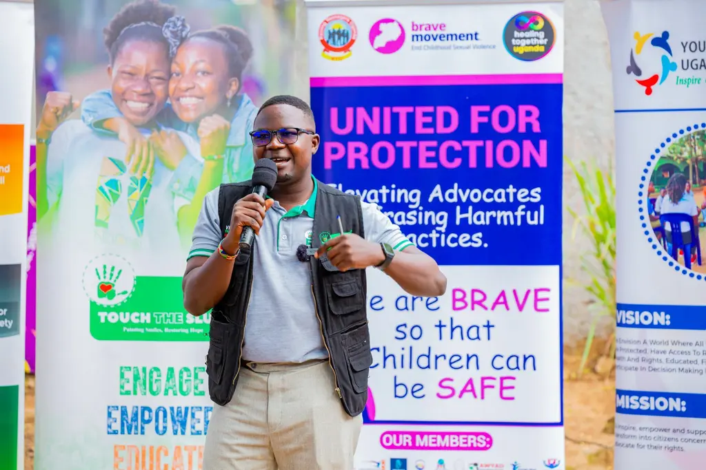 A man in casual clothing speaking into a microphone, standing in front of a colourful poster promoting child protection advocacy.
