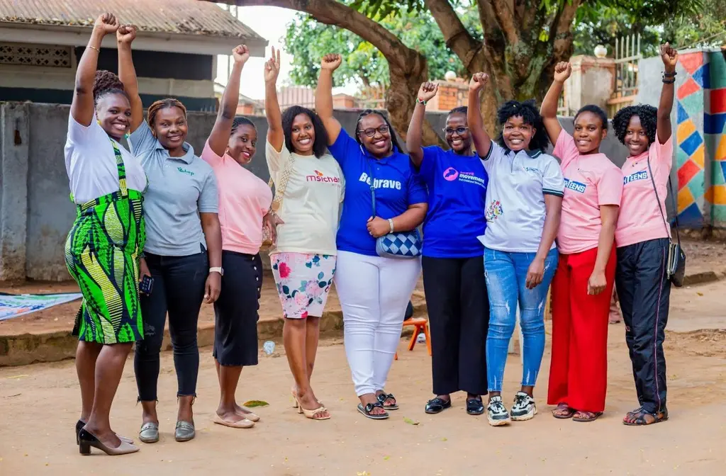 Group of diverse people, dressed casually, standing together with arms raised in celebration, surrounded by a colourful outdoor setting.