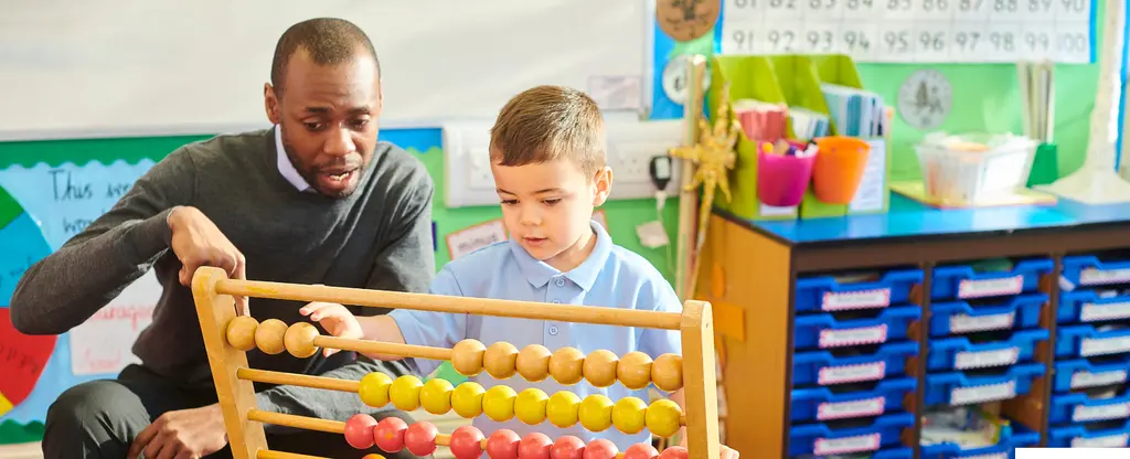A man and young boy using a colorful wooden abacus toy in a classroom setting with educational posters and storage units visible in the background.
