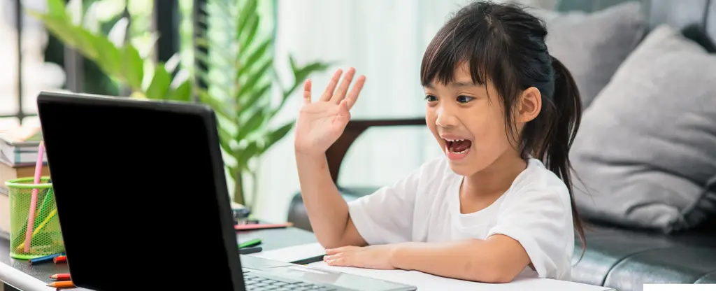 Young girl enthusiastically waving at laptop computer on desk, surrounded by plants and cushions.