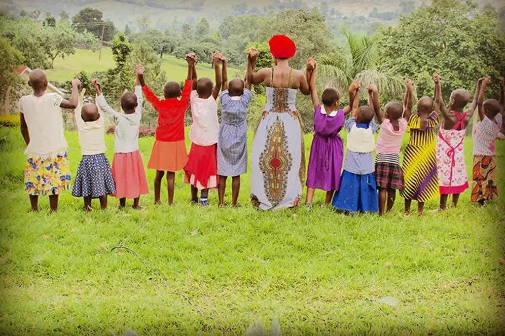 A large group of people, mostly children, stand in a field with their arms raised. They are wearing brightly colored traditional dress, including vibrant reds, purples, and yellows. The background features lush, verdant vegetation.