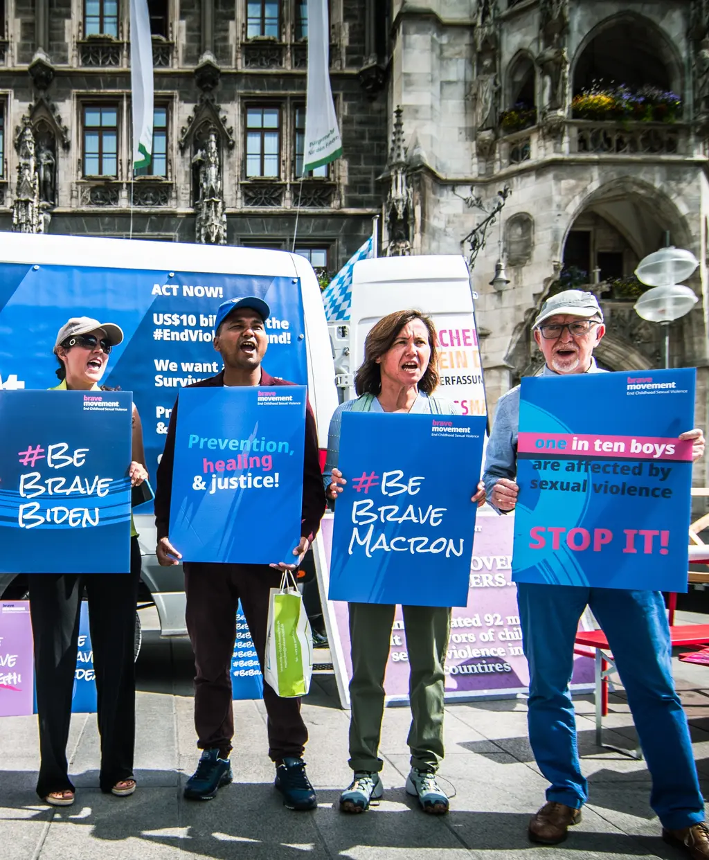 Protesters holding signs calling for action on Macron's policies, with slogans like "Be brave Macron" and "Stop it!" in front of a historic European building.