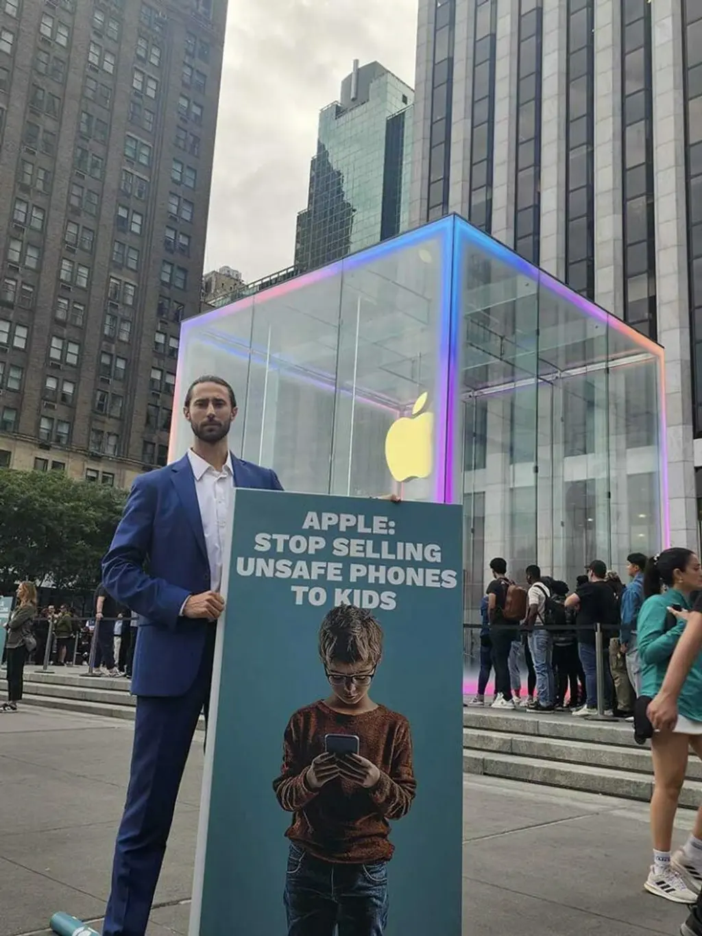 A man holding a sign that says "Apple: Stop selling unsafe phones to kids" in front of an Apple store with a glass cube structure and tall buildings in the background.