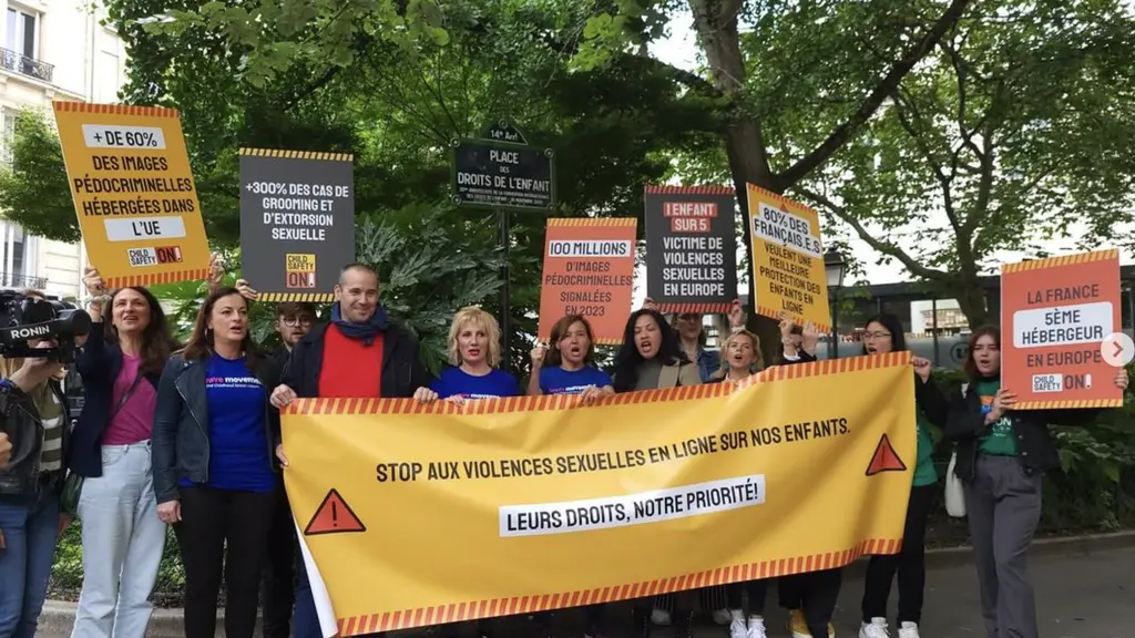 A group of people holding a large yellow banner that reads "STOP AUX VIOLENCES SEXUELLES EN LIGNE NOS ENFANTS, LEURS DROITS NOTRE PRIORITE" (Stop online sexual violence against our children, their rights are our priority). Protesters hold various placards and signs criticising online sexual violence against children.