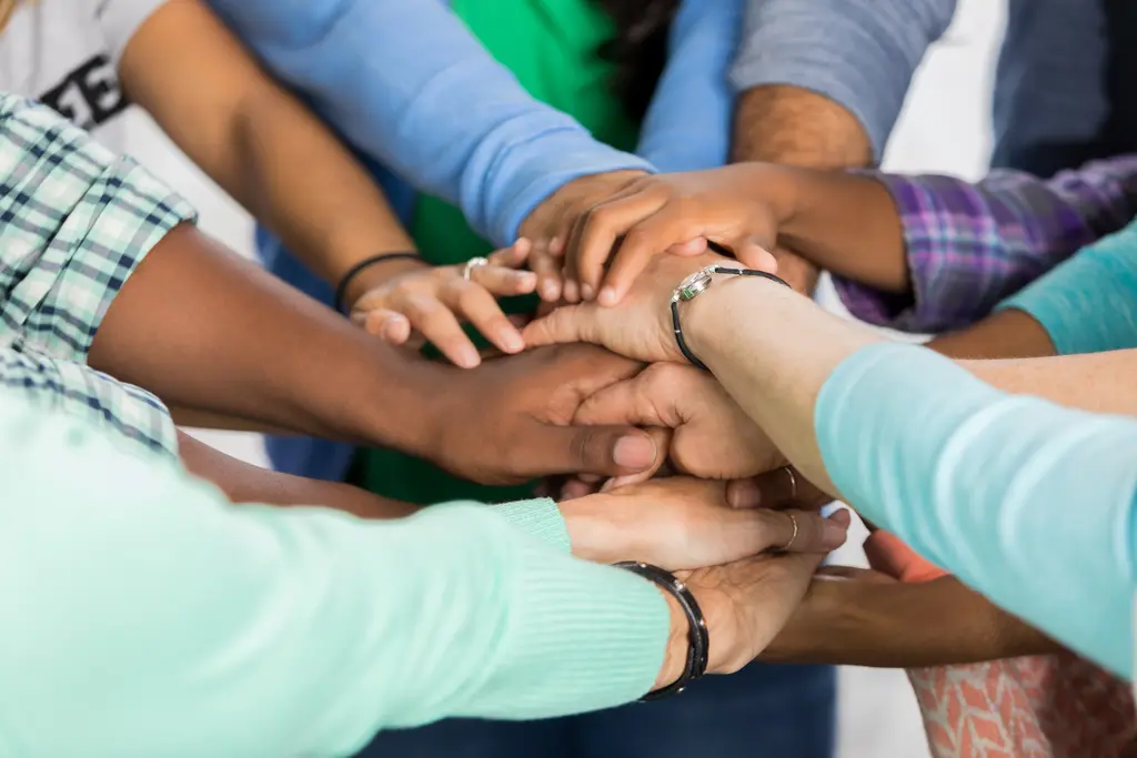 Diverse hands clasped together in unity and teamwork, various skin tones and bracelets visible.