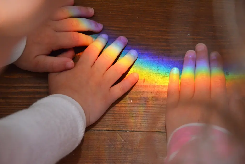 Hands with multicolored nails on wooden surface, creating a rainbow reflection.
