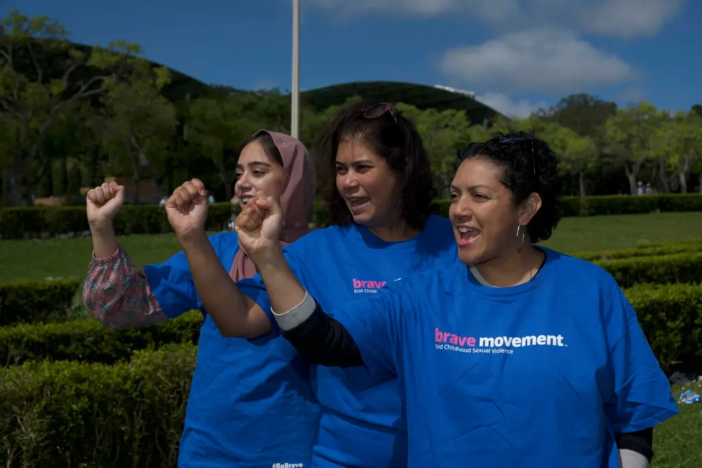 Three women wearing blue shirts with "Brave Movement" text, standing outdoors with arms raised, smiling.