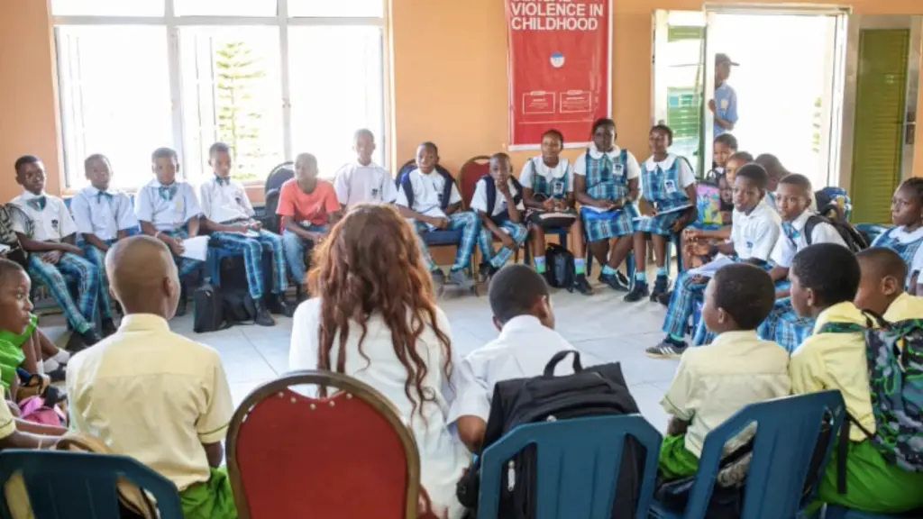 Group of children sitting in classroom, some in school uniforms. Walls have posters and signs.