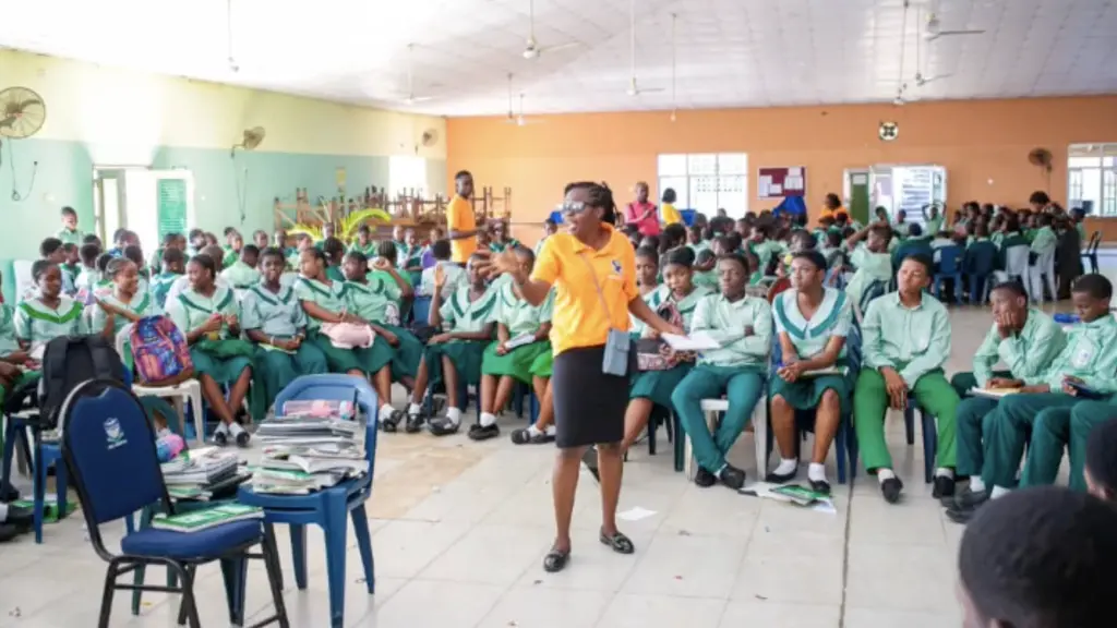 Large group of school children seated in a classroom, with a teacher standing in front of them.