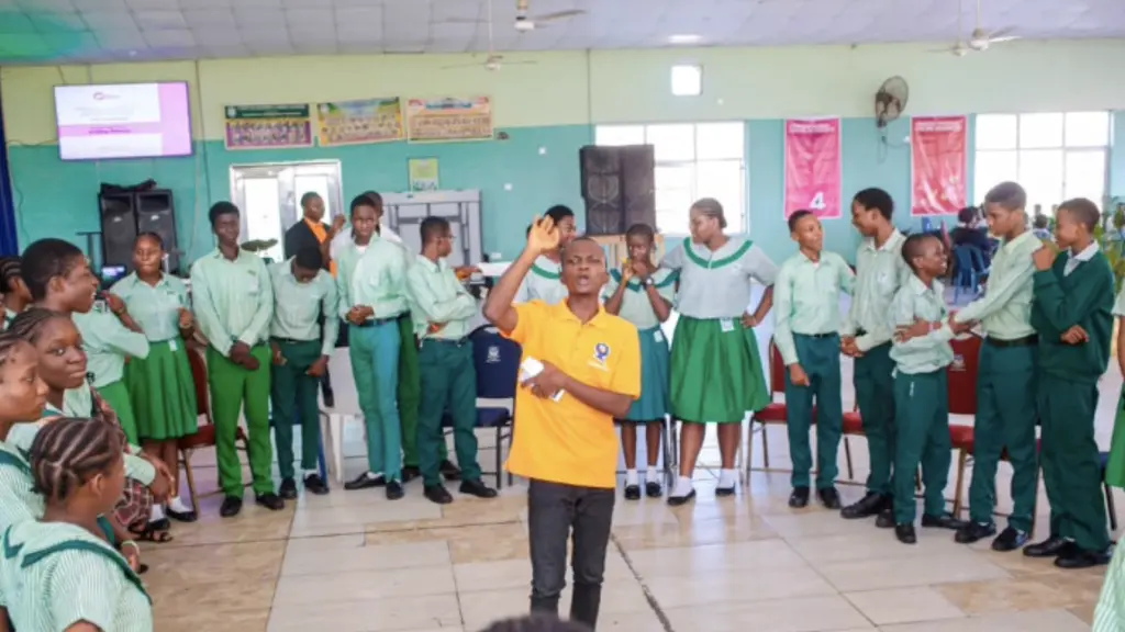 A group of children and adults in casual school uniforms, standing together in a classroom setting. There are various visual elements like chalkboards, desks, and other classroom furniture visible in the background.
