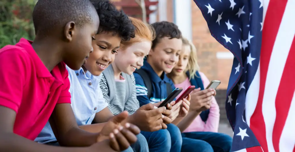 子供たちがスマートフォンを操作している様子。肌の色の異なる子供たちが集まり、フラグの背景の前でスマートフォンを見ている。