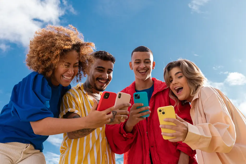 Four diverse young people laughing together while looking at their smartphones under a blue sky.